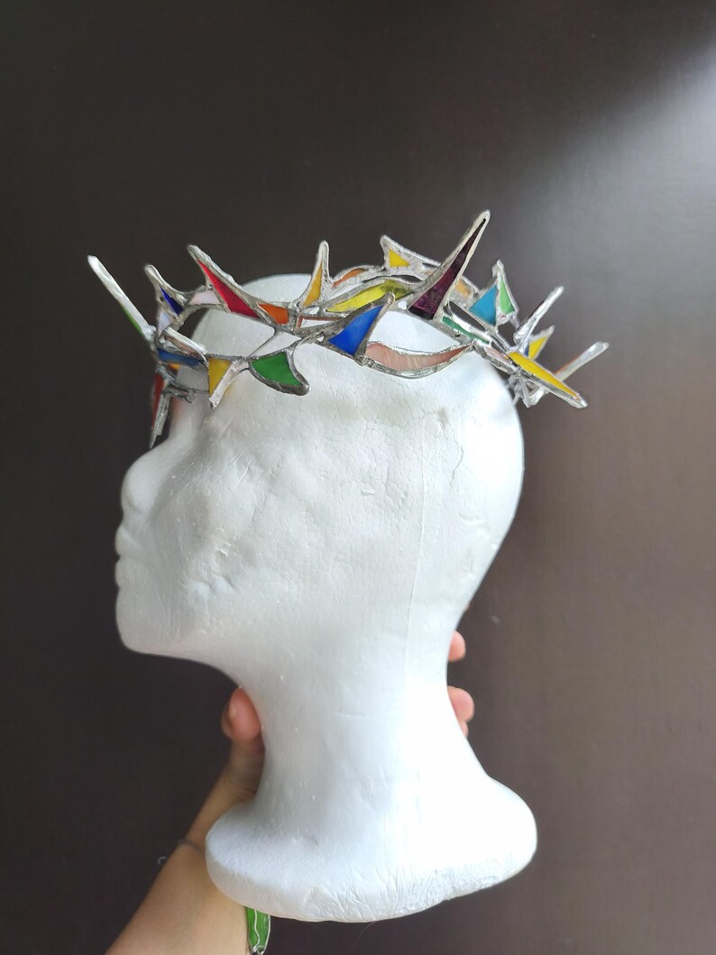 May include: A crown made of stained glass pieces in various colors including red, yellow, blue, green, and purple, joined together with silver-toned metal solder. The crown features sharp, pointed spikes and is displayed on a white mannequin head.