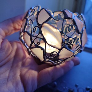 May include: A small, round, glass candle holder with a delicate wire frame and pink glass panels. The candle holder is lit and casting a warm glow.