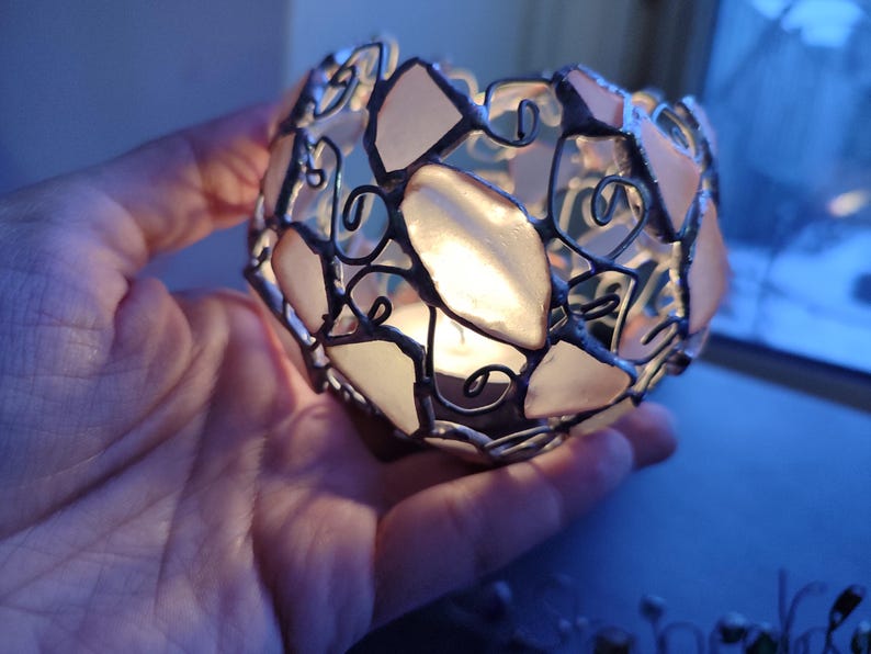 May include: A small, round, glass candle holder with a delicate wire frame and pink glass panels. The candle holder is lit and casting a warm glow.