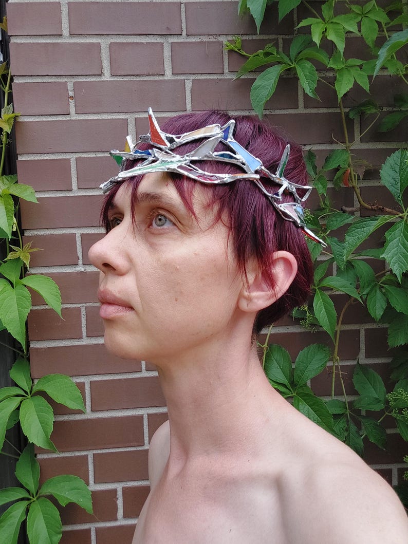 May include: A crown constructed from interconnected silver-toned metal branches and small, angular pieces of stained glass in various colors including red, blue, and white, designed to sit atop the head with a jagged, thorn-like appearance.