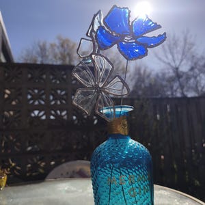 May include: A turquoise glass bottle with a textured surface, topped with a decorative floral arrangement made of blue and clear glass. The bottle has gold lettering. The arrangement is in front of a decorative fence and a bright sky.