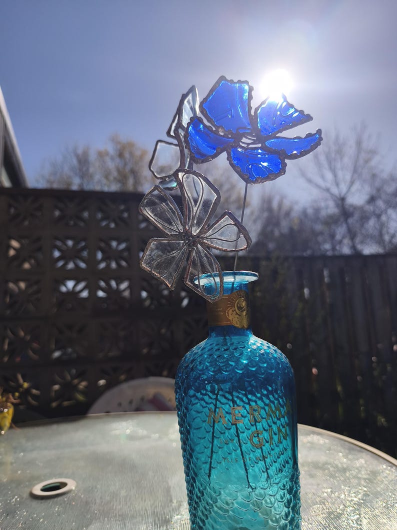 May include: A turquoise glass bottle with a textured surface, topped with a decorative floral arrangement made of blue and clear glass. The bottle has gold lettering. The arrangement is in front of a decorative fence and a bright sky.