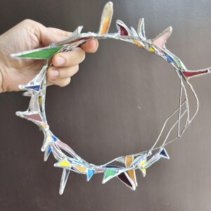 May include: A circular crown constructed from multiple small, triangular pieces of multicolored stained glass held together by silver-toned metal solder. The glass shards feature various hues including green, blue, yellow, orange, and red.