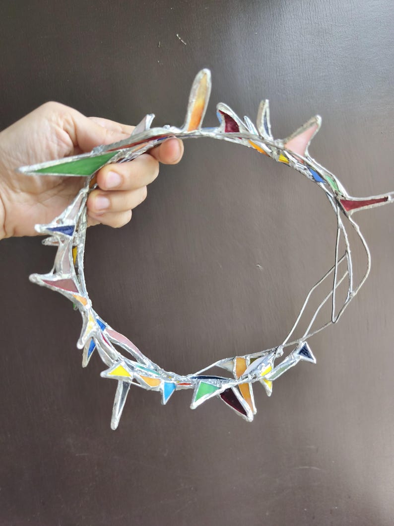 May include: A circular crown constructed from multiple small, triangular pieces of multicolored stained glass held together by silver-toned metal solder. The glass shards feature various hues including green, blue, yellow, orange, and red.