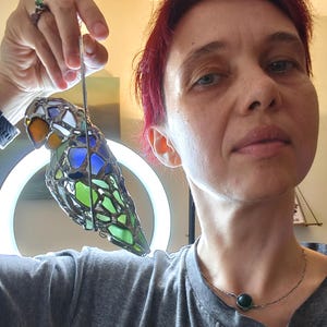 May include: A stained glass bird ornament with a colorful design. The bird features orange, blue, green, and white glass pieces, held together with metal. It is suspended by a thin wire, suitable for hanging. The bird is approximately 6 inches long.