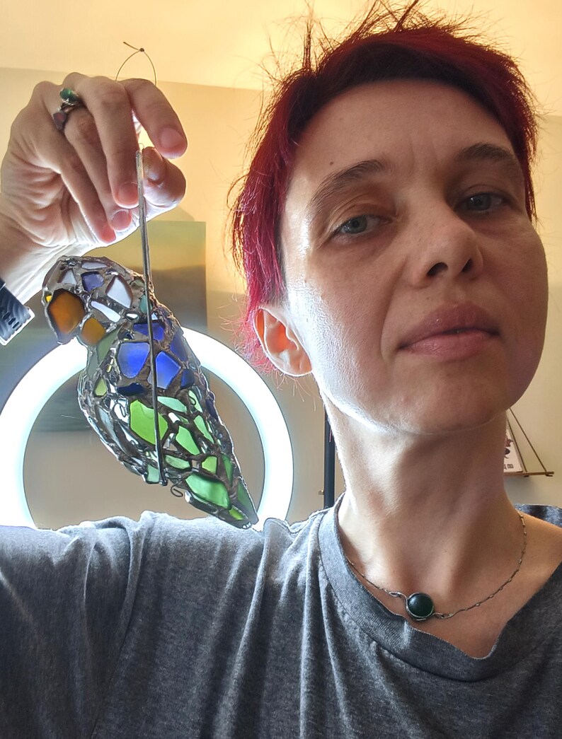 May include: A stained glass bird ornament with a colorful design. The bird features orange, blue, green, and white glass pieces, held together with metal. It is suspended by a thin wire, suitable for hanging. The bird is approximately 6 inches long.