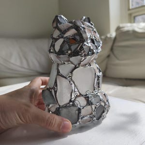 May include: A mosaic bear ornament made from white, grey, and brown glass pieces, held together with silver-coloured metal. The bear is in a sitting position, with its ears and body clearly defined.