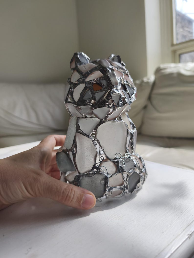 May include: A mosaic bear ornament made from white, grey, and brown glass pieces, held together with silver-coloured metal. The bear is in a sitting position, with its ears and body clearly defined.