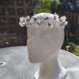 May include: A delicate floral crown featuring translucent, light pink and green flower accents. The flowers are attached to a silver-toned wire base. The crown is displayed on a white mannequin head, perfect for weddings or special occasions.