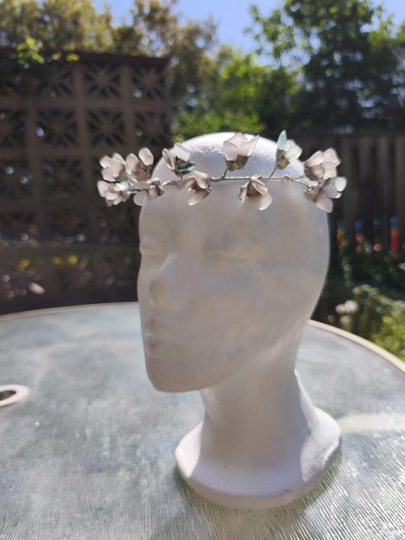 May include: A delicate floral crown featuring translucent, light pink and green flower accents. The flowers are attached to a silver-toned wire base. The crown is displayed on a white mannequin head, perfect for weddings or special occasions.
