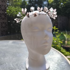 May include: A delicate floral crown with translucent, petal-shaped accents in shades of pink, gray, and clear. The flowers are attached to a thin, silver-colored wire. The crown is displayed on a white mannequin head, perfect for weddings, festivals, or special occasions.