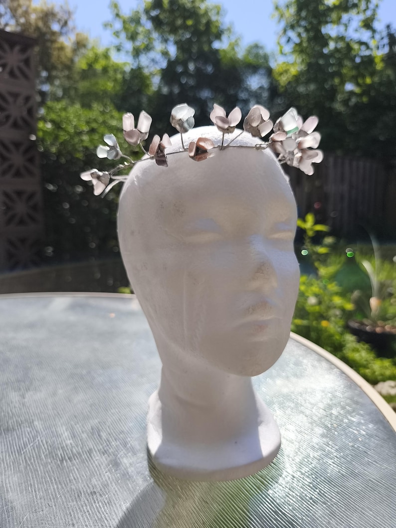 May include: A delicate floral crown with translucent, petal-shaped accents in shades of pink, gray, and clear. The flowers are attached to a thin, silver-colored wire. The crown is displayed on a white mannequin head, perfect for weddings, festivals, or special occasions.