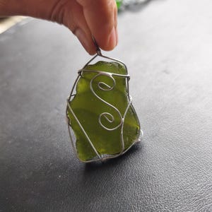 May include: A handcrafted pendant featuring a piece of sea glass in a vibrant green hue, encased in a silver wire frame. The wirework includes decorative swirls. The pendant is held up by a hand.