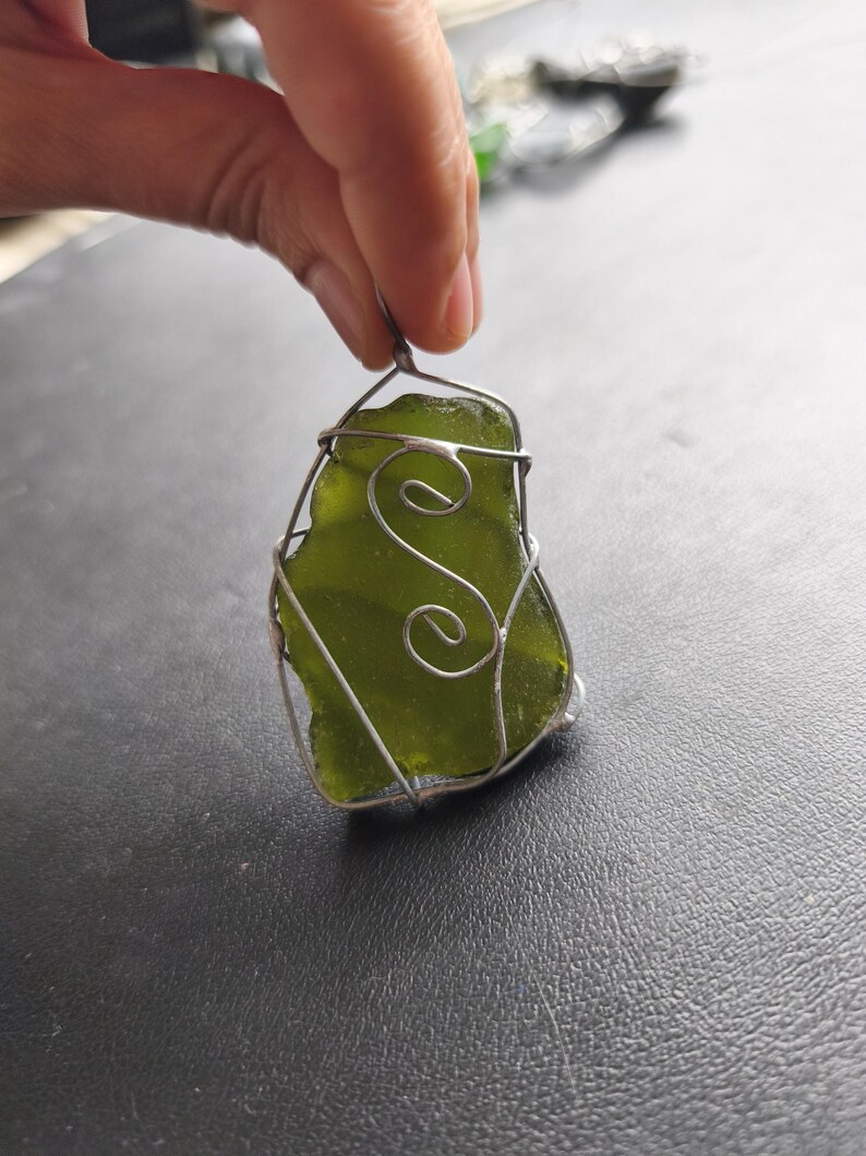 May include: A handcrafted pendant featuring a piece of sea glass in a vibrant green hue, encased in a silver wire frame. The wirework includes decorative swirls. The pendant is held up by a hand.