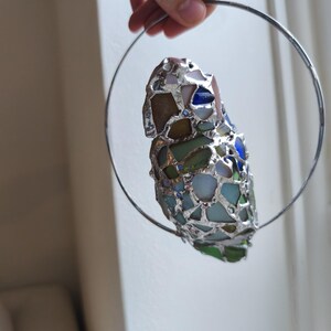 May include: A handcrafted stained glass ornament featuring a mosaic of colorful glass pieces in shades of blue, green, and brown, held within a silver metal ring. The ornament is suspended by a thin wire.