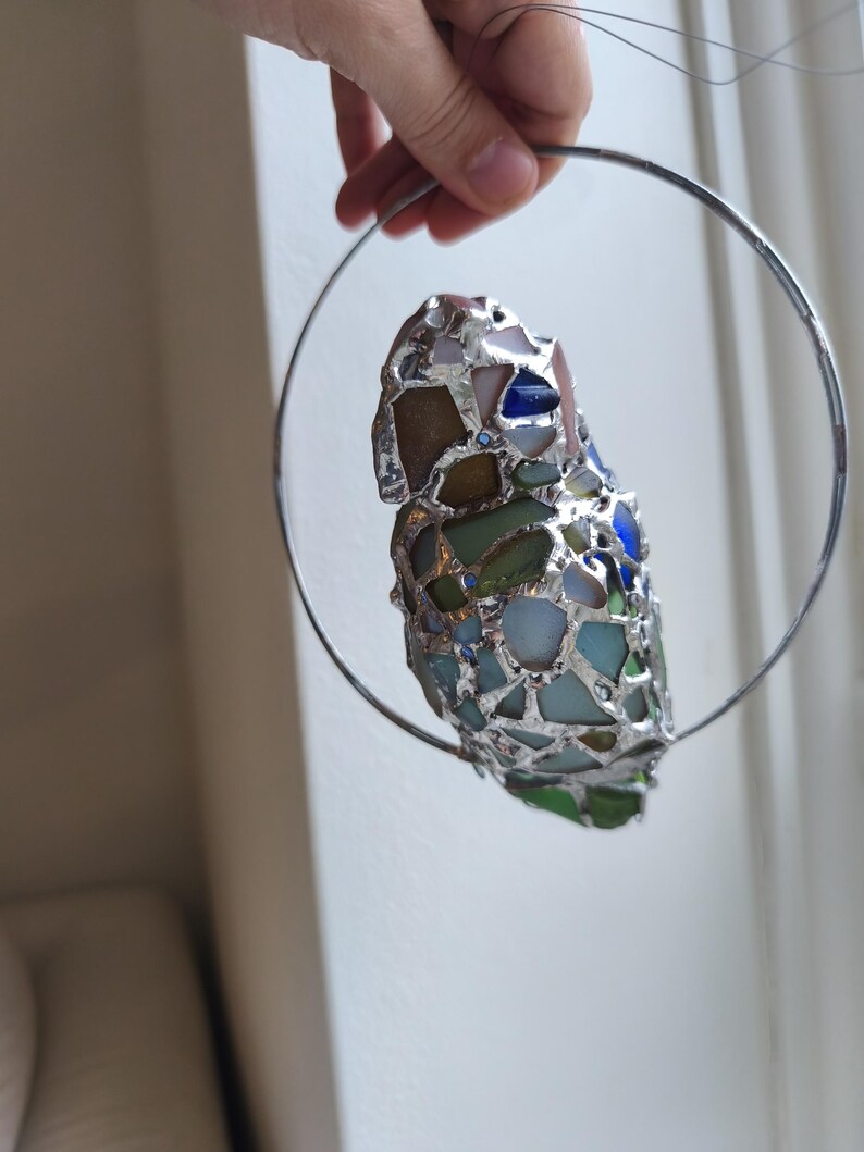 May include: A handcrafted stained glass ornament featuring a mosaic of colorful glass pieces in shades of blue, green, and brown, held within a silver metal ring. The ornament is suspended by a thin wire.