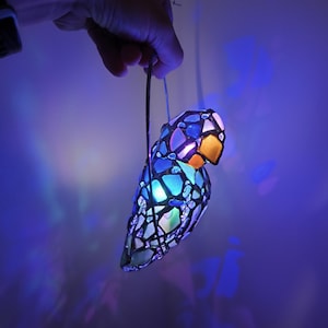 May include: A colorful, stained-glass parrot-shaped lamp with a glowing interior. The lamp features a mosaic design with blue, orange, and white glass pieces. It is suspended by a wire and emits a soft, ambient light, creating a decorative and artistic effect.