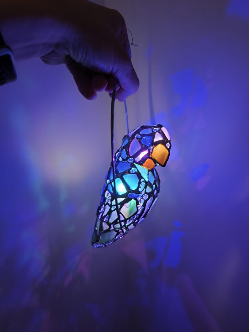 May include: A colorful, stained-glass parrot-shaped lamp with a glowing interior. The lamp features a mosaic design with blue, orange, and white glass pieces. It is suspended by a wire and emits a soft, ambient light, creating a decorative and artistic effect.