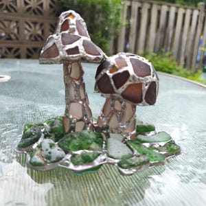 May include: A handcrafted glass art piece featuring two mushroom sculptures. The mushroom caps are made of brown and amber-colored glass pieces, while the stems are silver. The base is adorned with green sea glass and silver accents.