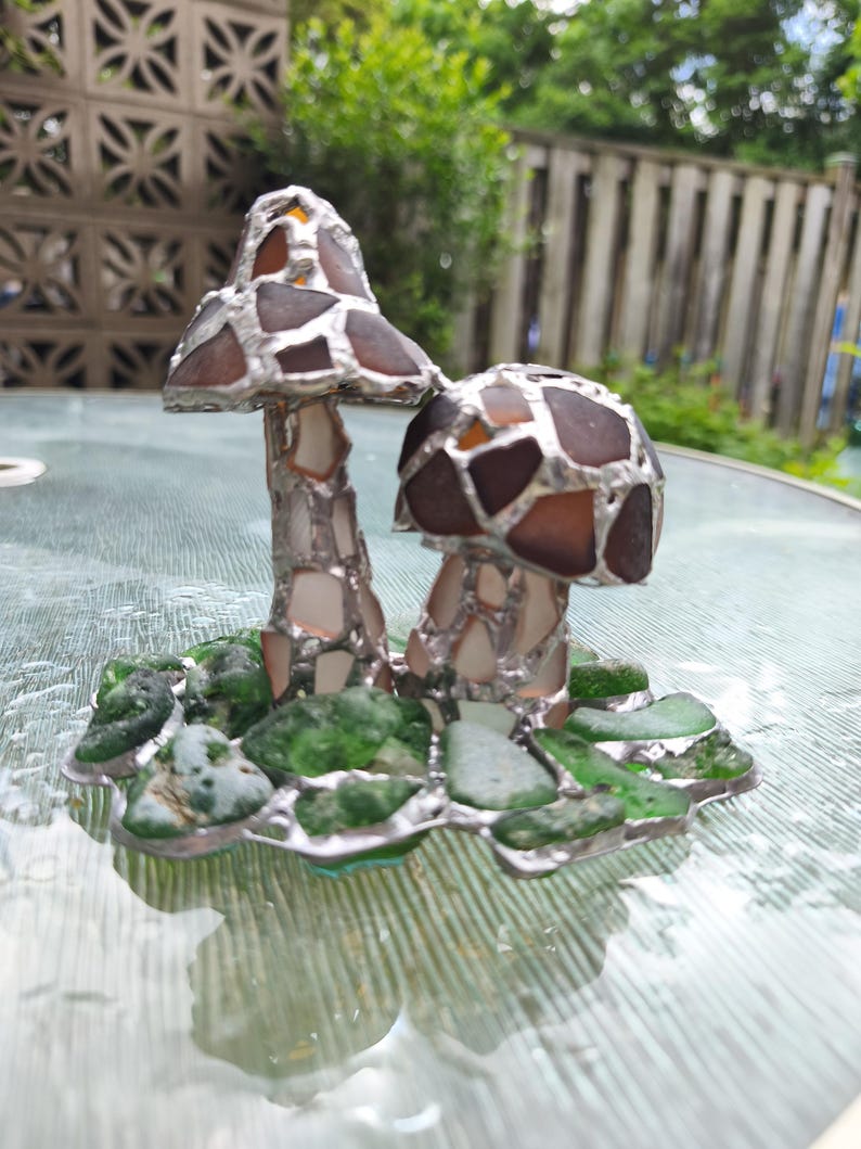 May include: A handcrafted glass art piece featuring two mushroom sculptures. The mushroom caps are made of brown and amber-colored glass pieces, while the stems are silver. The base is adorned with green sea glass and silver accents.