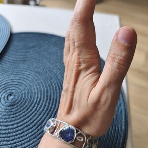 May include: A silver bracelet with a series of irregularly shaped, deep blue stones. The bracelet is worn on a wrist, with the hand in the background. The bracelet's design features a delicate, openwork pattern. The stones are set within the silver frame.
