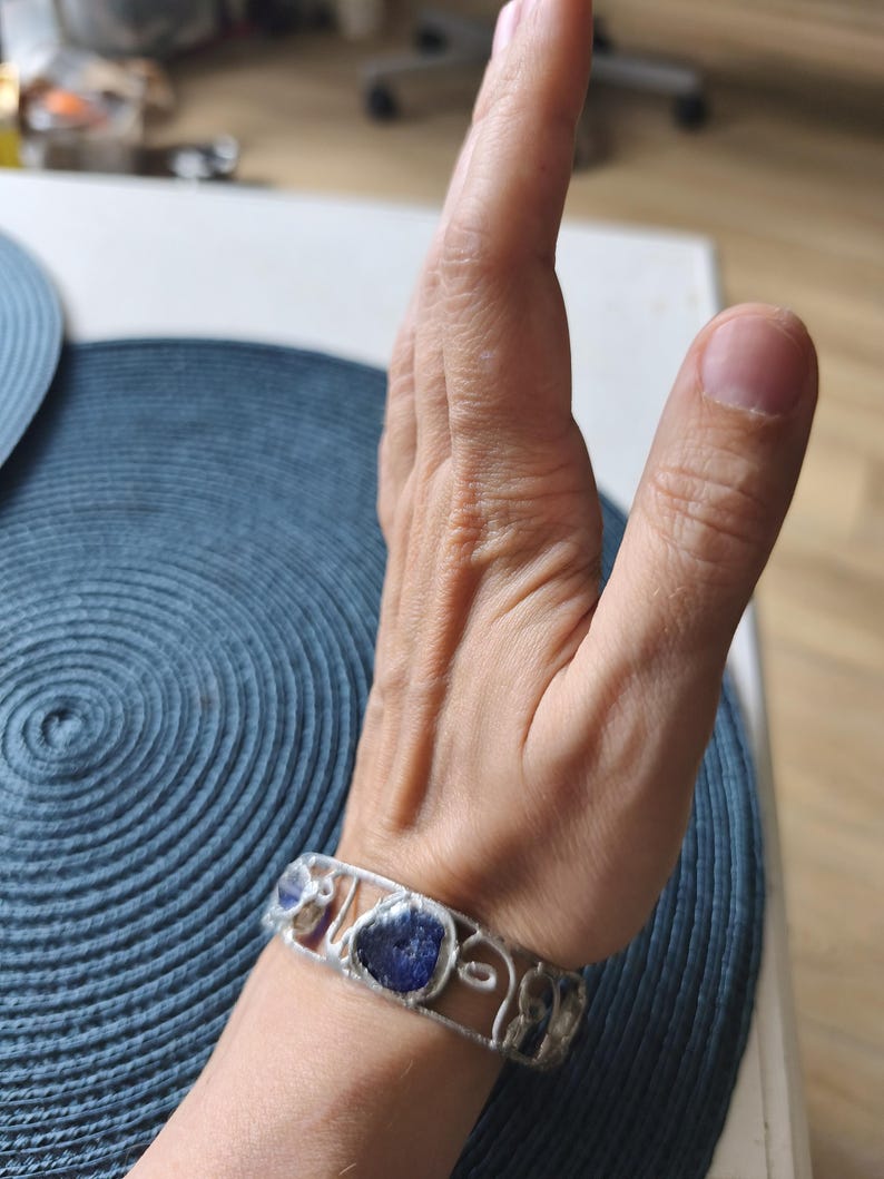 May include: A silver bracelet with a series of irregularly shaped, deep blue stones. The bracelet is worn on a wrist, with the hand in the background. The bracelet's design features a delicate, openwork pattern. The stones are set within the silver frame.