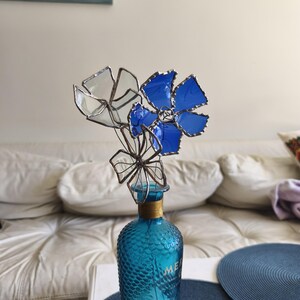 May include: A turquoise glass bottle with a textured surface, adorned with a cluster of stained glass flowers in blue and clear. The flowers have a metallic trim, and the bottle has a gold-colored neck.