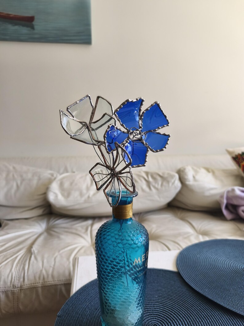May include: A turquoise glass bottle with a textured surface, adorned with a cluster of stained glass flowers in blue and clear. The flowers have a metallic trim, and the bottle has a gold-colored neck.