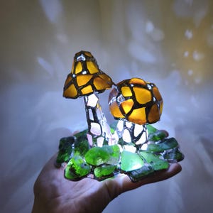 May include: A handcrafted mushroom lamp with a stained glass design. The lamp features two mushroom caps in shades of amber and brown, with a mosaic pattern. The base is made of green glass pieces. The lamp is lit from within, creating a warm glow.