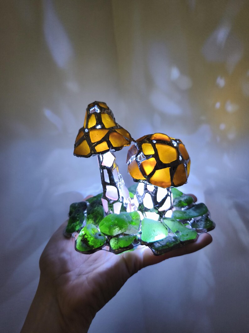 May include: A handcrafted mushroom lamp with a stained glass design. The lamp features two mushroom caps in shades of amber and brown, with a mosaic pattern. The base is made of green glass pieces. The lamp is lit from within, creating a warm glow.