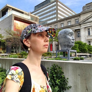 May include: A colorful baseball cap with a geometric design. The cap features a mix of black, white, and vibrant colors. The person is wearing a floral top and a black backpack. The background includes buildings and a sculpture.