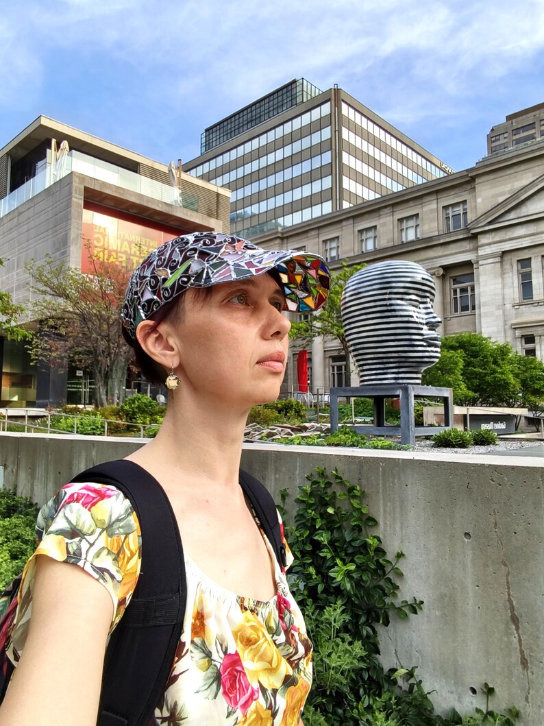 May include: A colorful baseball cap with a geometric design. The cap features a mix of black, white, and vibrant colors. The person is wearing a floral top and a black backpack. The background includes buildings and a sculpture.