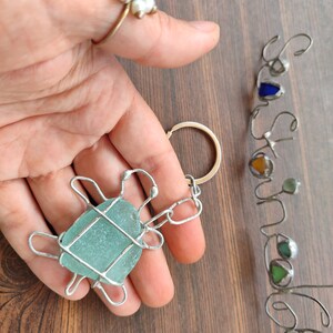 K&ouml;nnte beinhalten: Ein Schl&uuml;sselanh&auml;nger aus Silberdraht in Form einer Schildkr&ouml;te mit einem hellblauen St&uuml;ck Seetang in der Mitte. Der Schl&uuml;sselanh&auml;nger wird in einer Hand mit einem Perlring am Zeigefinger gehalten.