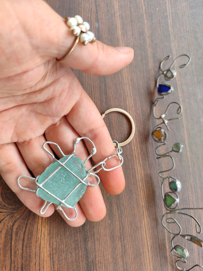 K&ouml;nnte beinhalten: Ein Schl&uuml;sselanh&auml;nger aus Silberdraht in Form einer Schildkr&ouml;te mit einem hellblauen St&uuml;ck Seetang in der Mitte. Der Schl&uuml;sselanh&auml;nger wird in einer Hand mit einem Perlring am Zeigefinger gehalten.