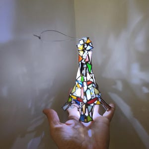 May include: A stained glass Eiffel Tower ornament in a variety of colours. The ornament is made of small pieces of glass, including red, yellow, green, and blue. It has a wire loop for hanging. The ornament is held in a hand.