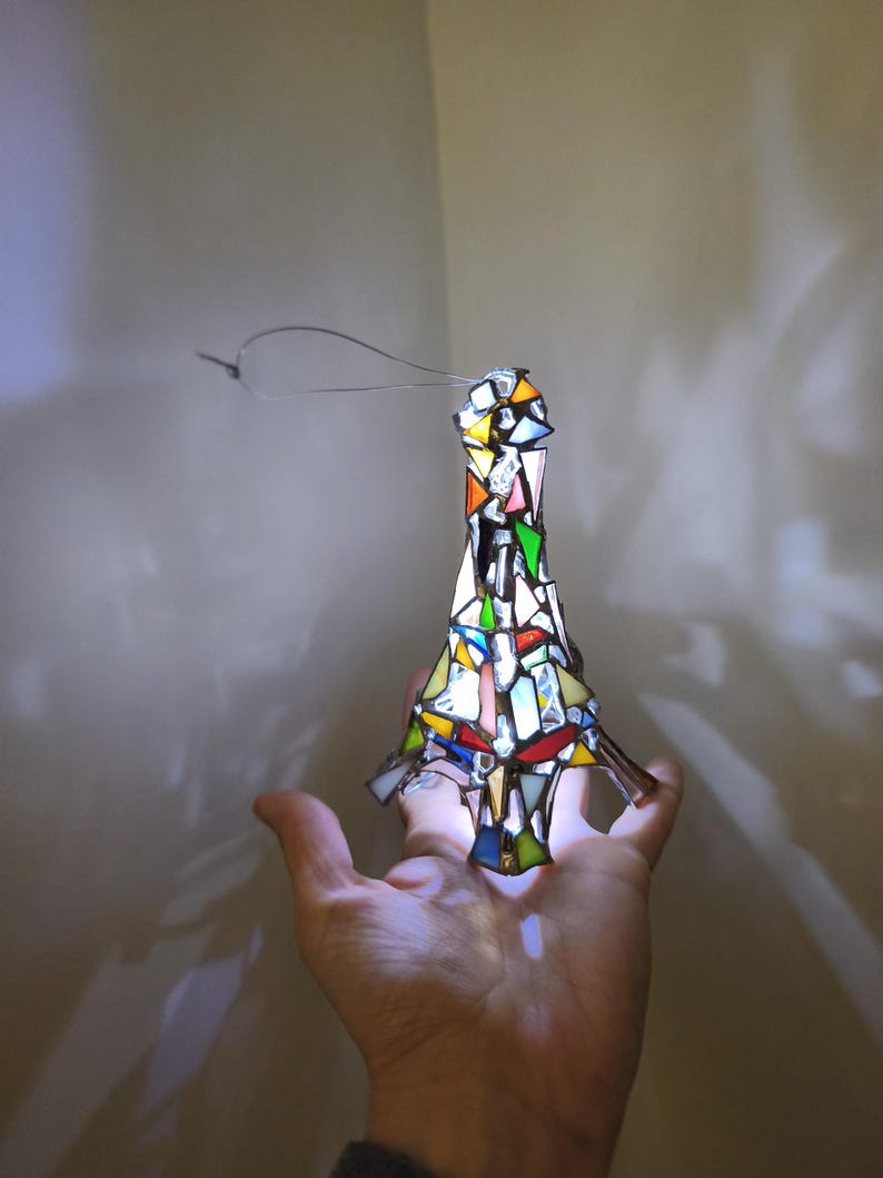 May include: A stained glass Eiffel Tower ornament in a variety of colours. The ornament is made of small pieces of glass, including red, yellow, green, and blue. It has a wire loop for hanging. The ornament is held in a hand.