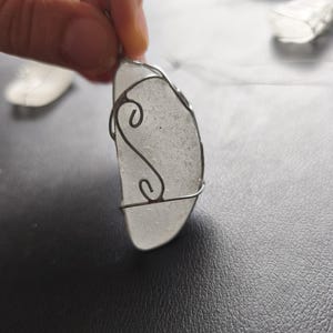 May include: A handcrafted pendant featuring a piece of frosted sea glass, encased in a silver wire frame. The wire is shaped into decorative swirls. The pendant hangs from a thin, clear cord, suitable for wearing as a necklace.