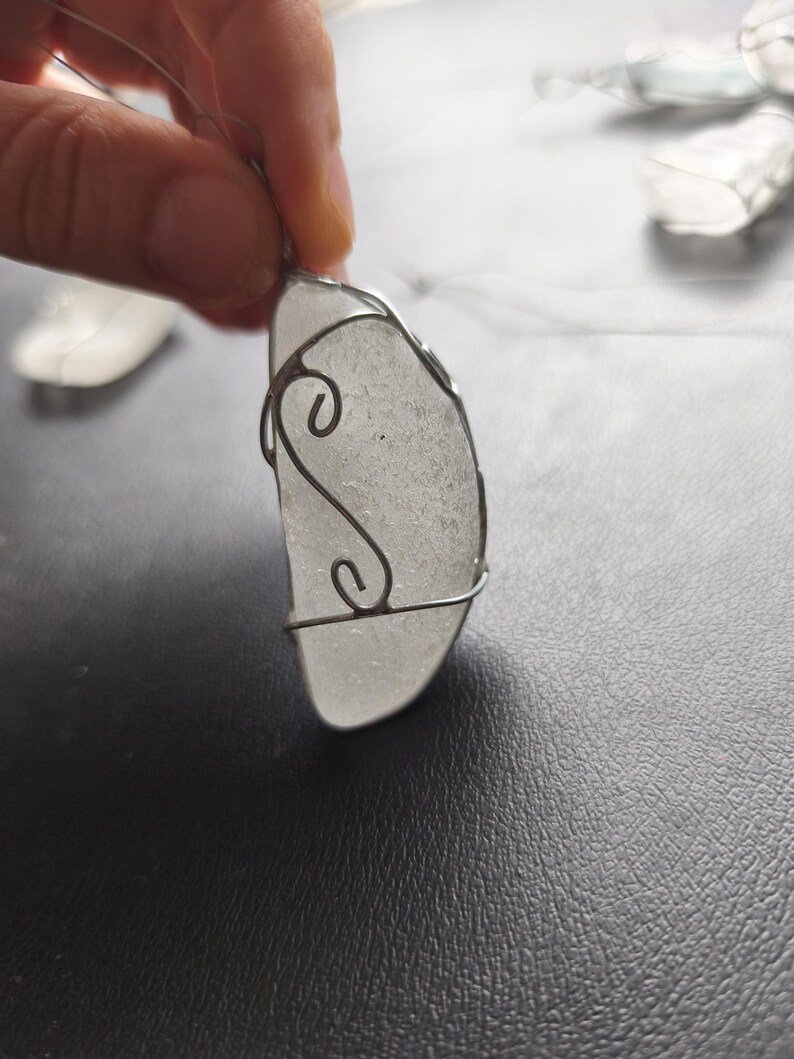 May include: A handcrafted pendant featuring a piece of frosted sea glass, encased in a silver wire frame. The wire is shaped into decorative swirls. The pendant hangs from a thin, clear cord, suitable for wearing as a necklace.