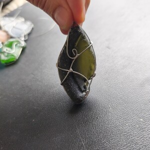 May include: A teardrop-shaped pendant made from dark green sea glass, wrapped in silver-colored wire. The pendant is held up against a dark surface. Other pieces of sea glass jewelry are visible in the background.