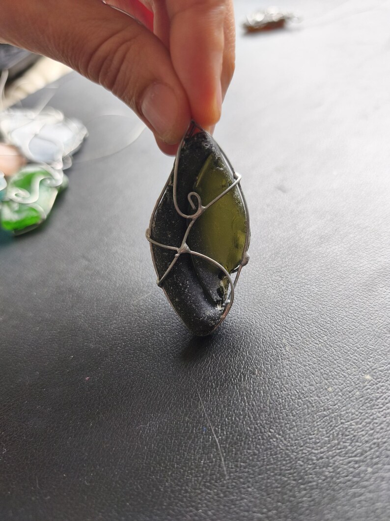 May include: A teardrop-shaped pendant made from dark green sea glass, wrapped in silver-colored wire. The pendant is held up against a dark surface. Other pieces of sea glass jewelry are visible in the background.