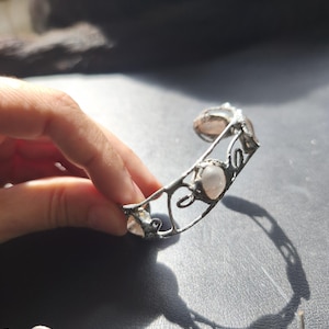 May include: A silver-toned cuff bracelet with three light pink, oval-shaped stones. The bracelet has an open design with decorative swirls and is held by a hand against a dark background. The bracelet is a piece of jewelry.