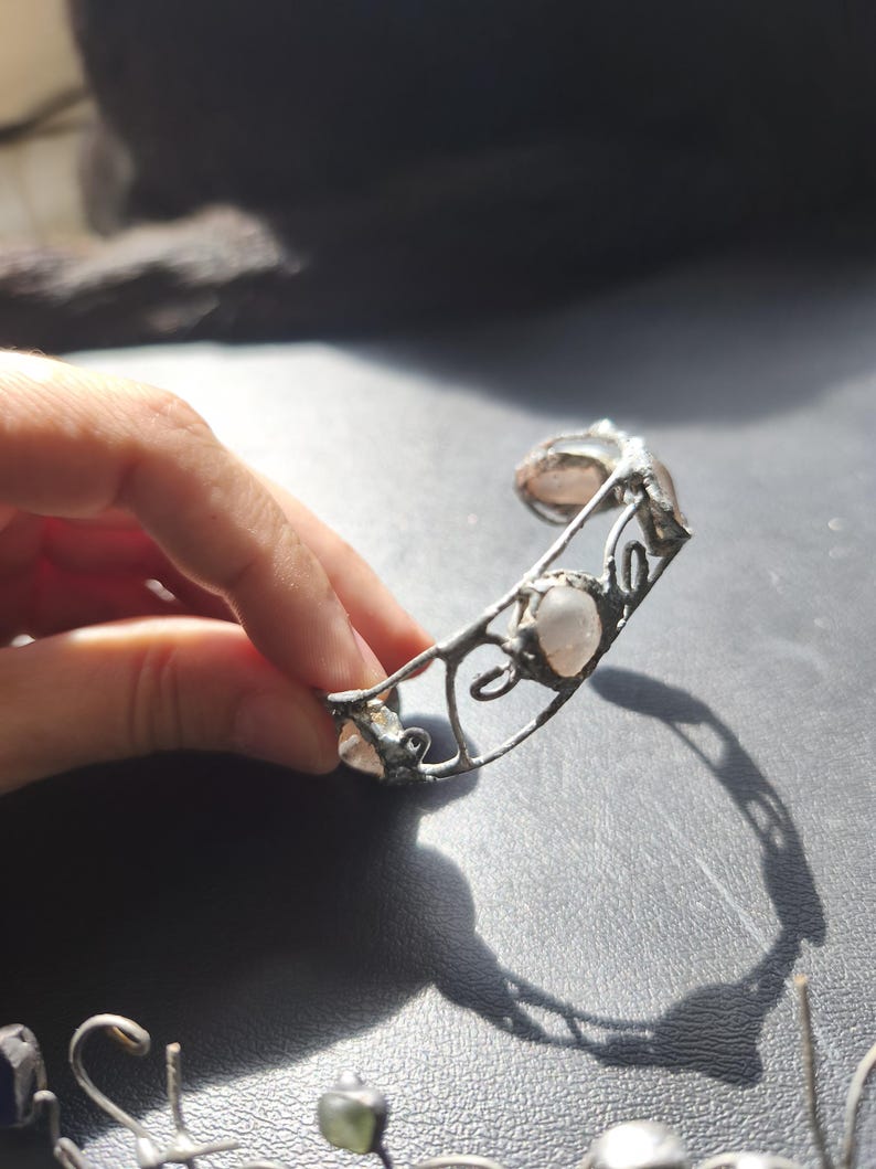 May include: A silver-toned cuff bracelet with three light pink, oval-shaped stones. The bracelet has an open design with decorative swirls and is held by a hand against a dark background. The bracelet is a piece of jewelry.