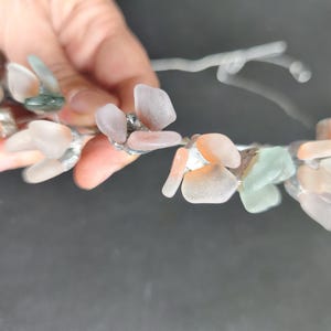 May include: A delicate floral crown featuring clusters of pastel-colored sea glass pieces. The sea glass is in shades of pink, peach, and green, attached to a silver-colored wire base. The crown is held against a dark background.