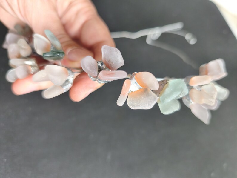 May include: A delicate floral crown featuring clusters of pastel-colored sea glass pieces. The sea glass is in shades of pink, peach, and green, attached to a silver-colored wire base. The crown is held against a dark background.