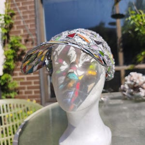 May include: A decorative hat with a visor, featuring a mosaic design of colorful glass pieces set in a silver metal frame. The hat is displayed on a white mannequin head, reflecting sunlight.