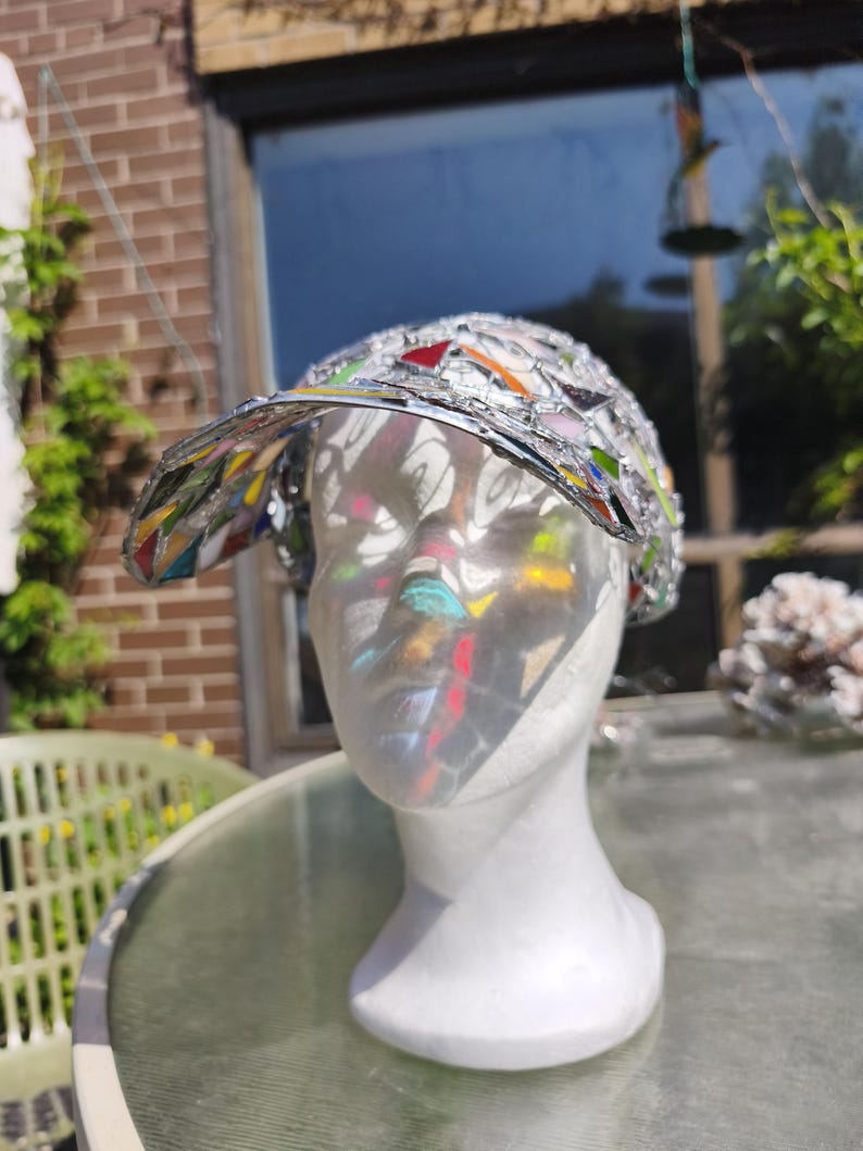 May include: A decorative hat with a visor, featuring a mosaic design of colorful glass pieces set in a silver metal frame. The hat is displayed on a white mannequin head, reflecting sunlight.