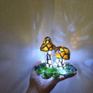 May include: A decorative mushroom lamp with two mushroom caps in shades of yellow and orange, with a mosaic-like pattern. The stems are white with black accents, and the base is green. The lamp is lit, creating a warm glow.