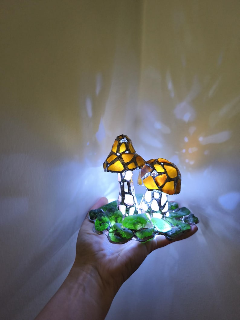 May include: A decorative mushroom lamp with two mushroom caps in shades of yellow and orange, with a mosaic-like pattern. The stems are white with black accents, and the base is green. The lamp is lit, creating a warm glow.