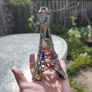 May include: A stained glass Eiffel Tower ornament. The tower is made with silver-coloured metal and features various coloured glass pieces, including red, blue, yellow, and green. A silver string is attached for hanging. The ornament is held in a hand.