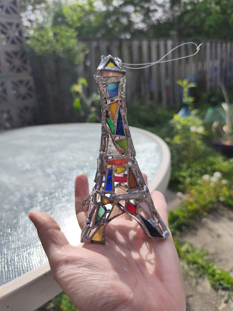 May include: A stained glass Eiffel Tower ornament. The tower is made with silver-coloured metal and features various coloured glass pieces, including red, blue, yellow, and green. A silver string is attached for hanging. The ornament is held in a hand.
